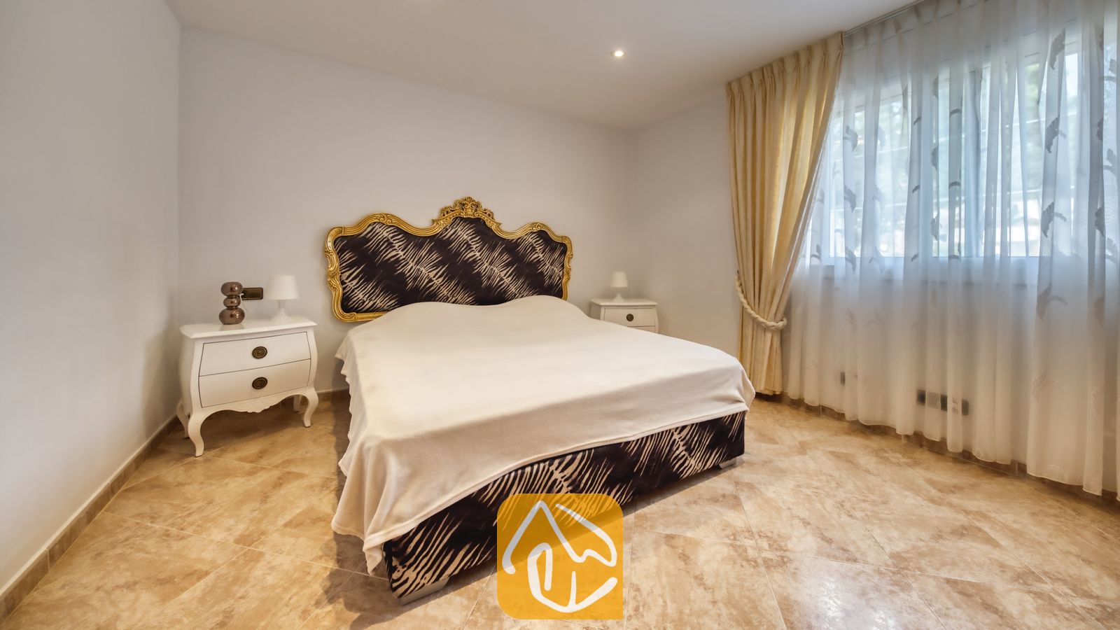 Vakantievilla's Costa Brava Spanje - Villa Lorena - Slaapkamer met een groot tweepersoonsbed met een sierlijk, goudomrand hoofdbord in een zebrasterkte, witte nachtkastjes en inbetween gordijnen voor het raam.