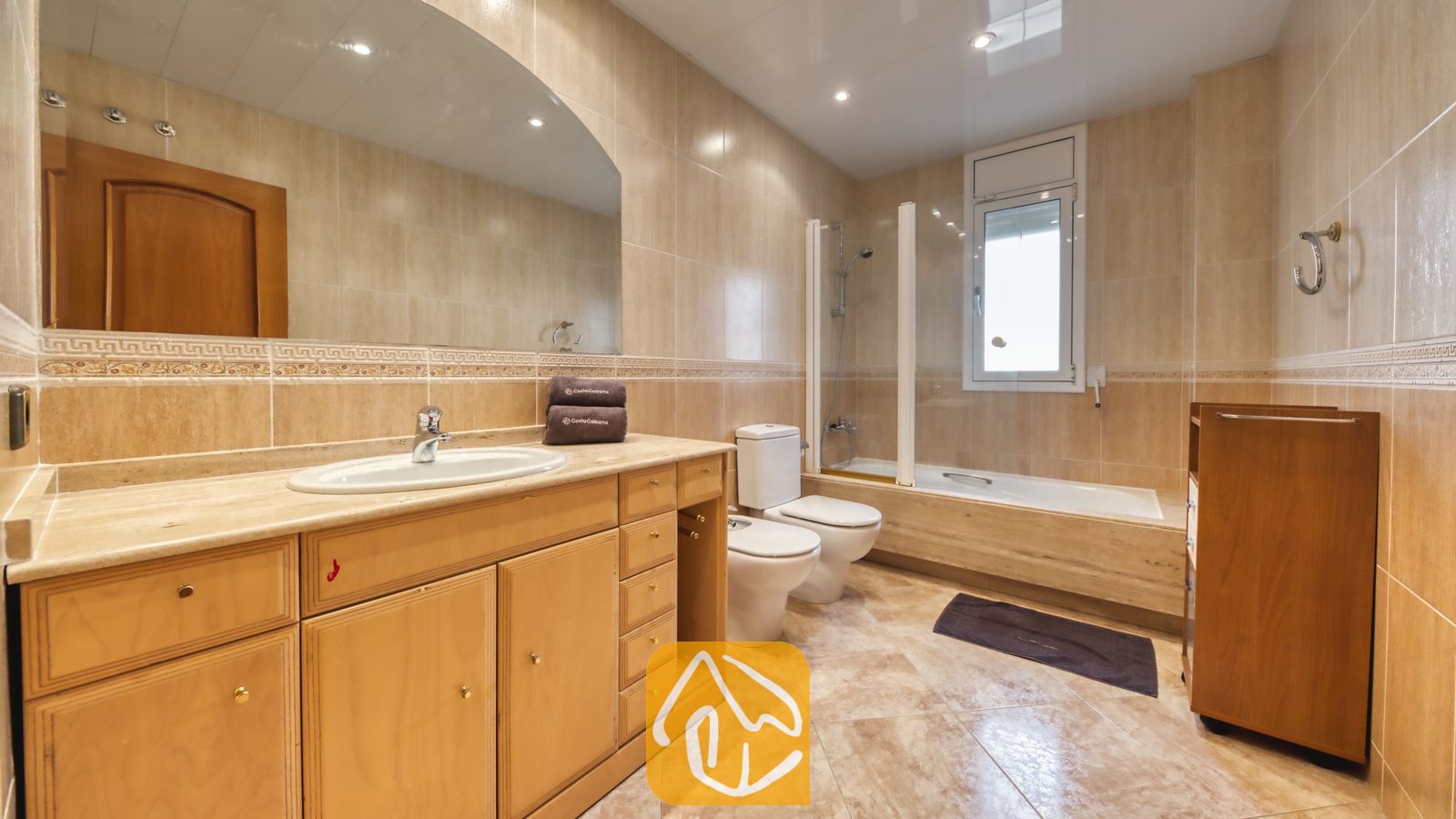 Vakantievilla's Costa Brava Spanje - Villa Lorena - Ruime badkamer met beige tegelwerk, voorzien van een grote houten wastafelmeubel met geïntegreerde wasbak, een brede wandspiegel, een bidet en een ligbad met glazen douchescherm onder een raam.