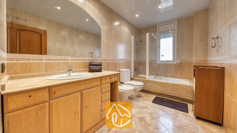 Villas de vacances Costa Brava Espagne - Villa Lorena - Spacieuse salle de bain carrelée de beige, comprenant un grand meuble sous-vasque en bois avec vasque intégrée, un large miroir mural, un bidet et une baignoire dotée d'une paroi de douche en verre, située sous une fenêtre.
