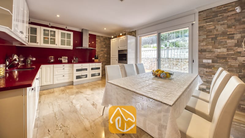Vakantievilla's Costa Brava Spanje - Villa Lorena - Keuken en eethoek in Villa Lorena met witte kasten, rode aanrechtbladen, een accentmuur van steen en een eettafel met witte lederen stoelen.