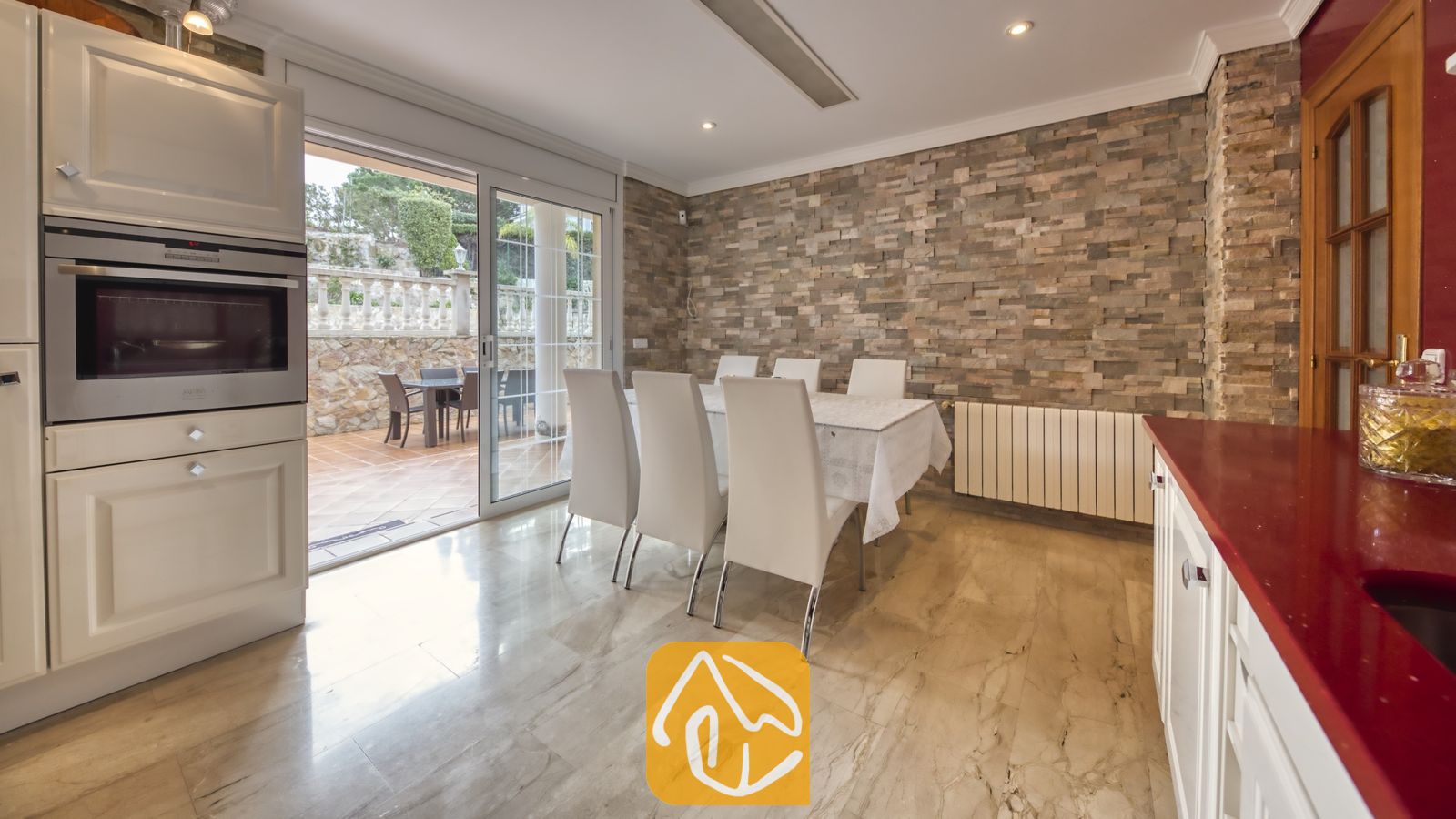 Vakantievilla's Costa Brava Spanje - Villa Lorena - Eethoek binnen in Villa Lorena met een witte tafel en zes witte stoelen, naast een stenen accentmuur en glazen schuifdeuren die uitkomen op een buitenterras.