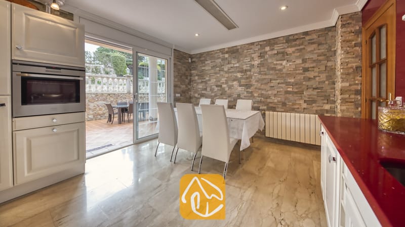 Villas de vacances Costa Brava Espagne - Villa Lorena - Espace repas à l'intérieur de la Villa Lorena, doté d'une table blanche et de six chaises blanches, situé près d'un mur d'accent en pierre et de portes coulissantes en verre s'ouvrant sur un patio extérieur.