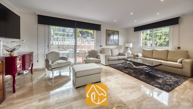 Vakantievilla's Costa Brava Spanje - Villa Lorena - Ruime woonkamer in Villa Lorena, voorzien van beige leren banken, een glazen salontafel op een donker vloerkleed en glazen schuifdeuren die toegang bieden tot een terras.