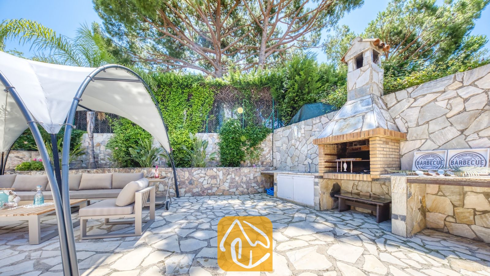 Vakantievilla's Costa Brava Spanje - Villa Lorena - Buitenbarbecuezone bij Villa Lorena, voorzien van een stenen grill, een terras met zitgedeelte onder een witte luifel en een bestrating van natuursteen.