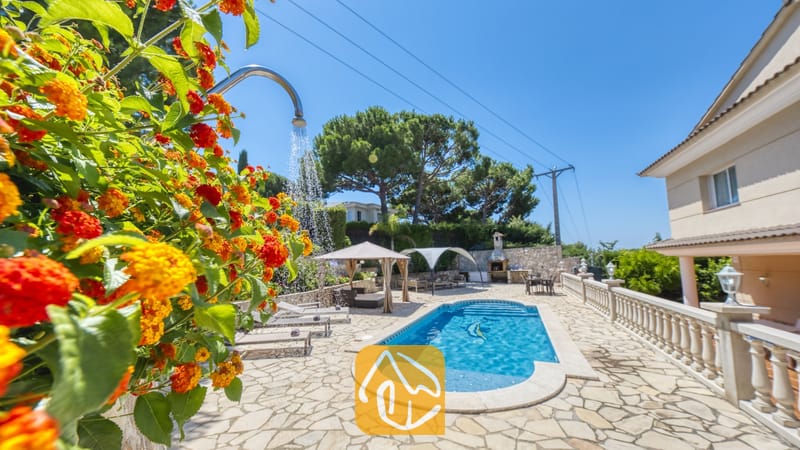 Vakantievilla's Costa Brava Spanje - Villa Lorena - Het buitenzwembad van Villa Lorena, met een blauw zwembad, een stenen terras, ligstoelen en oranje bloemen op de voorgrond.