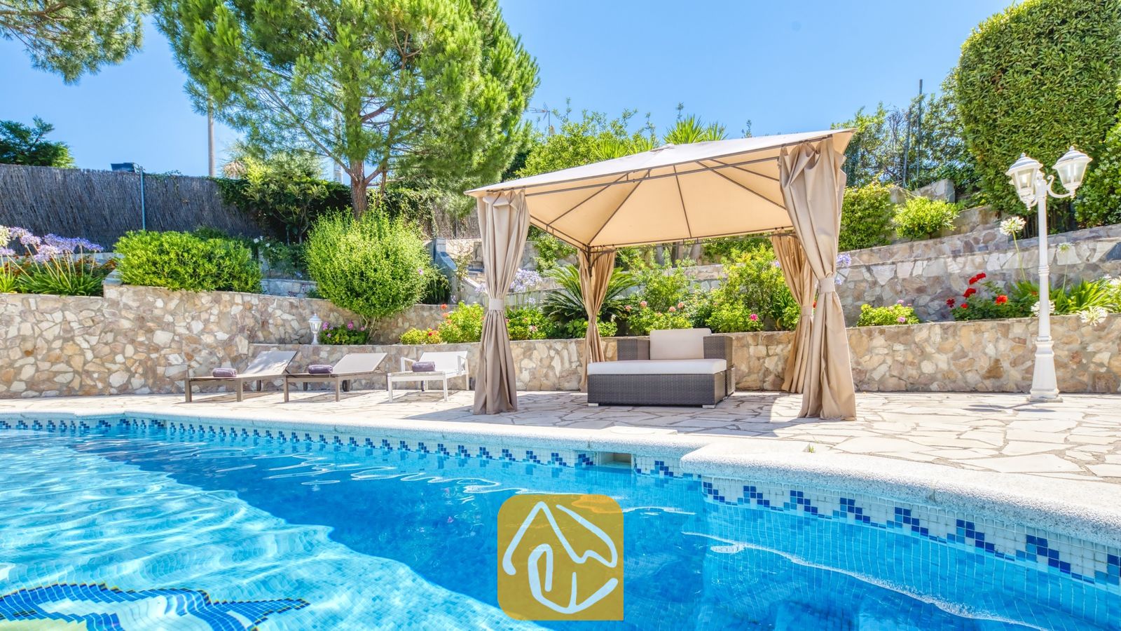 Vakantievilla's Costa Brava Spanje - Villa Lorena - Privézwembad bij Villa Lorena met blauw betegelde randen, een beige stoffen prieel over het buitenzitje, stenen keermuren en omringend groen.