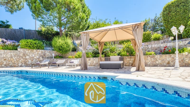 Vakantievilla's Costa Brava Spanje - Villa Lorena - Privézwembad bij Villa Lorena met blauw betegelde randen, een beige stoffen prieel over het buitenzitje, stenen keermuren en omringend groen.
