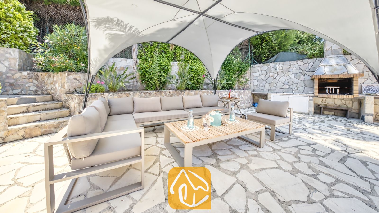 Vakantievilla's Costa Brava Spanje - Villa Lorena - Buitenlounge onder een witte overkapping bij Villa Lorena, voorzien van een grijze hoekbank, een houten salontafel en een stenen BBQ-openhaard.