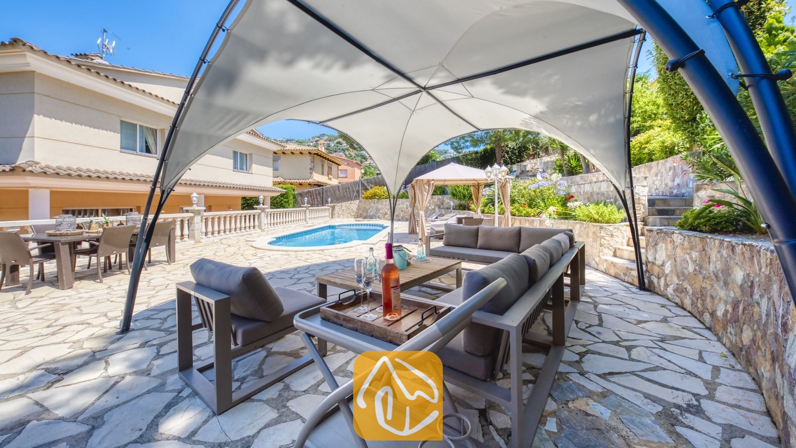 Vakantievilla's Costa Brava Spanje - Villa Lorena - Overdekte buitenlounge met grijs meubilair onder een witte overkapping bij Villa Lorena, met uitzicht op het zwembad en de patio.