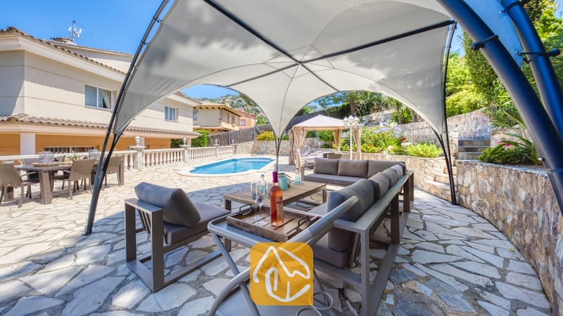 Vakantievilla's Costa Brava Spanje - Villa Lorena - Overdekte buitenlounge met grijs meubilair onder een witte overkapping bij Villa Lorena, met uitzicht op het zwembad en de patio.