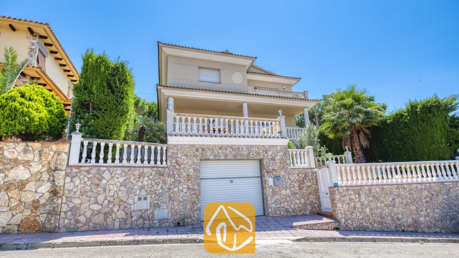 Vakantievilla's Costa Brava Spanje - Villa Lorena - Buitengevel van Villa Lorena, waarop een woning met meerdere verdiepingen te zien is, voorzien van een stenen sokkel, garagedeur, witte balustrades en omringende palmbomen tegen een blauwe lucht.