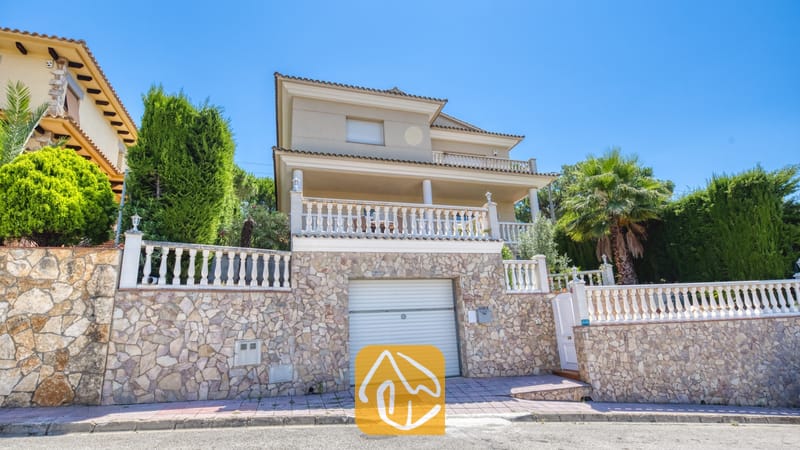 Villas de vacances Costa Brava Espagne - Villa Lorena - Façade extérieure de la Villa Lorena présentant une maison à paliers multiples dotée d'un mur de soubassement en pierre, d'une porte de garage, de balustrades blanches et de palmiers environnants sous un ciel bleu.