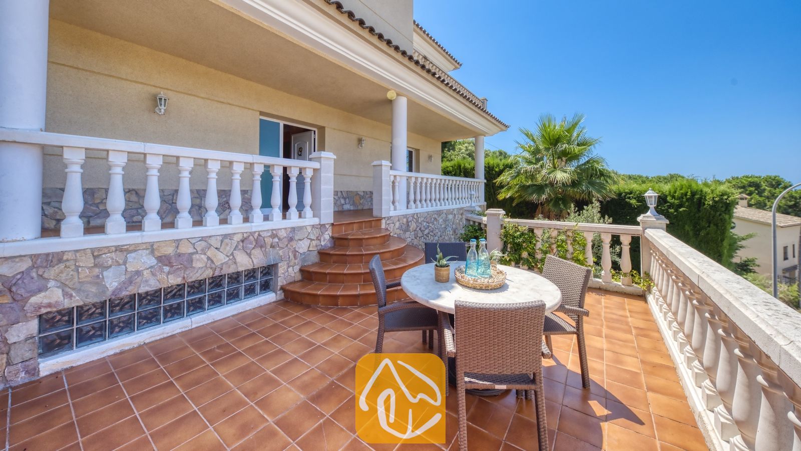 Vakantievilla's Costa Brava Spanje - Villa Lorena - Zonnig buitenterras bij Villa Lorena, voorzien van een ronde eettafel met gevlochten stoelen, terracotta vloertegels en witte balustrades tegen een helderblauwe hemel.