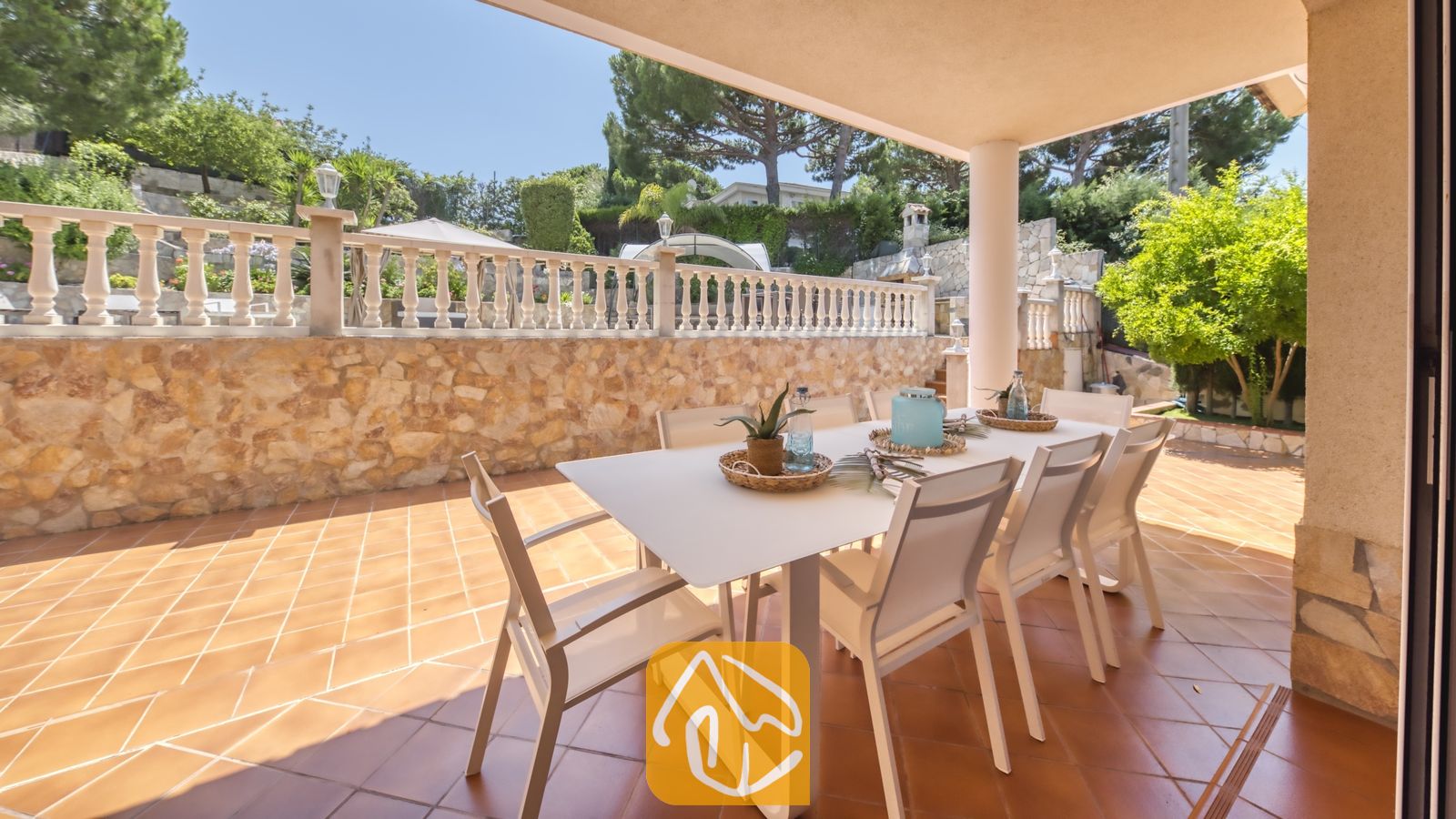 Vakantievilla's Costa Brava Spanje - Villa Lorena - Overdekt terras bij Villa Lorena met een witte eettafel omringd door bijpassende stoelen op een tegelvloer, met een stenen keermuur en een decoratieve balustrade op de achtergrond.