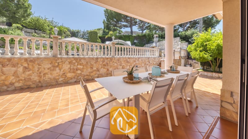 Vakantievilla's Costa Brava Spanje - Villa Lorena - Overdekt terras bij Villa Lorena met een witte eettafel omringd door bijpassende stoelen op een tegelvloer, met een stenen keermuur en een decoratieve balustrade op de achtergrond.