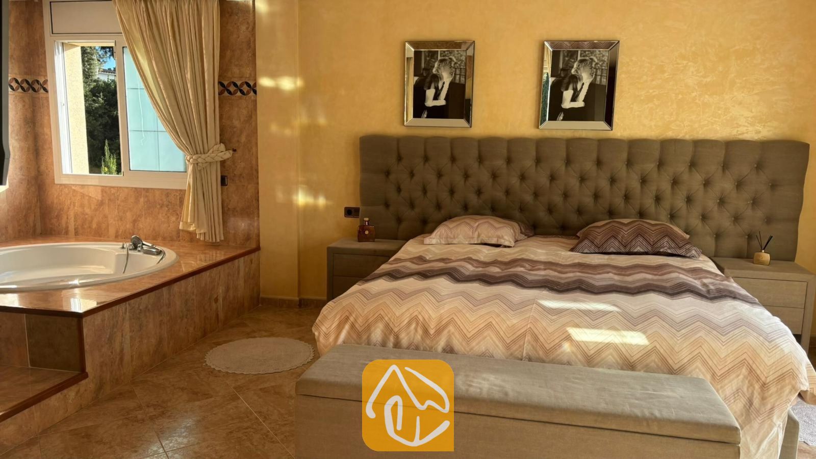 Vakantievilla's Costa Brava Spanje - Villa Lorena - Slaapkamer met een groot gecapitonneerd hoofdbord, beddengoed met patroon en een geïntegreerd hoekbad naast het raam.