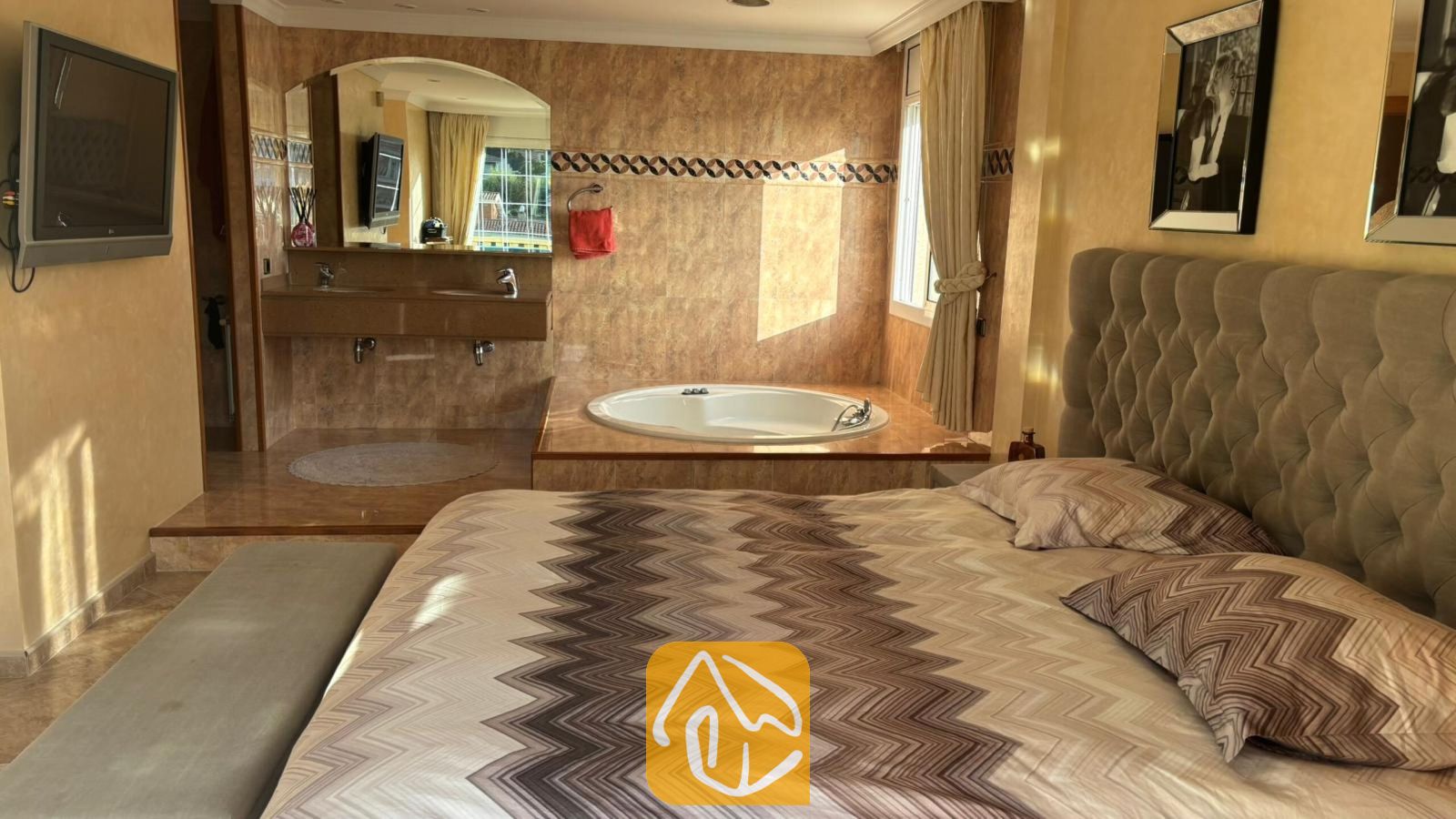 Vakantievilla's Costa Brava Spanje - Villa Lorena - Slaapkamer met een gestoffeerd hoofdbord en bed voorzien van gedessineerd beddengoed, een geïntegreerde open badkamer met een rond ligbad en betegelde muren.