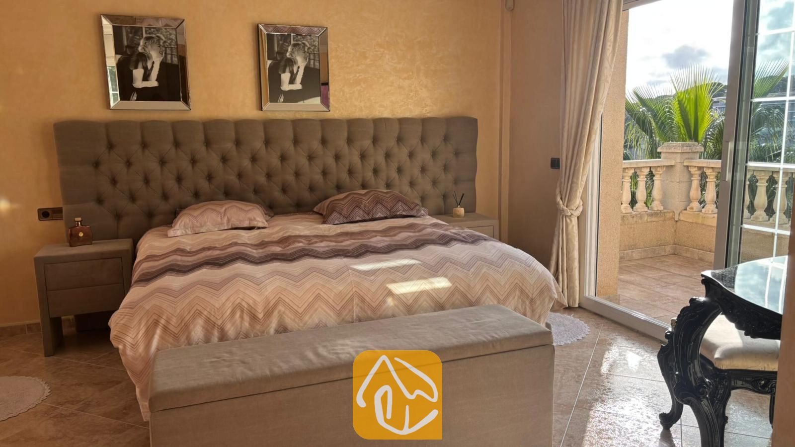 Vakantievilla's Costa Brava Spanje - Villa Lorena - Slaapkamer met een groot gecapitonneerd grijs hoofdbord, beddengoed met patroon, bijpassende nachtkastjes en een schuifpui die uitkomt op een balkon met palmbomen.
