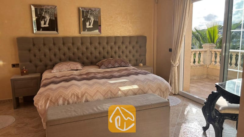 Villas de vacances Costa Brava Espagne - Villa Lorena - Chambre avec une grande tête de lit capitonnée en tissu gris, une literie à motifs, des tables de chevet assorties et des portes coulissantes en verre s'ouvrant sur un balcon bordé de palmiers.