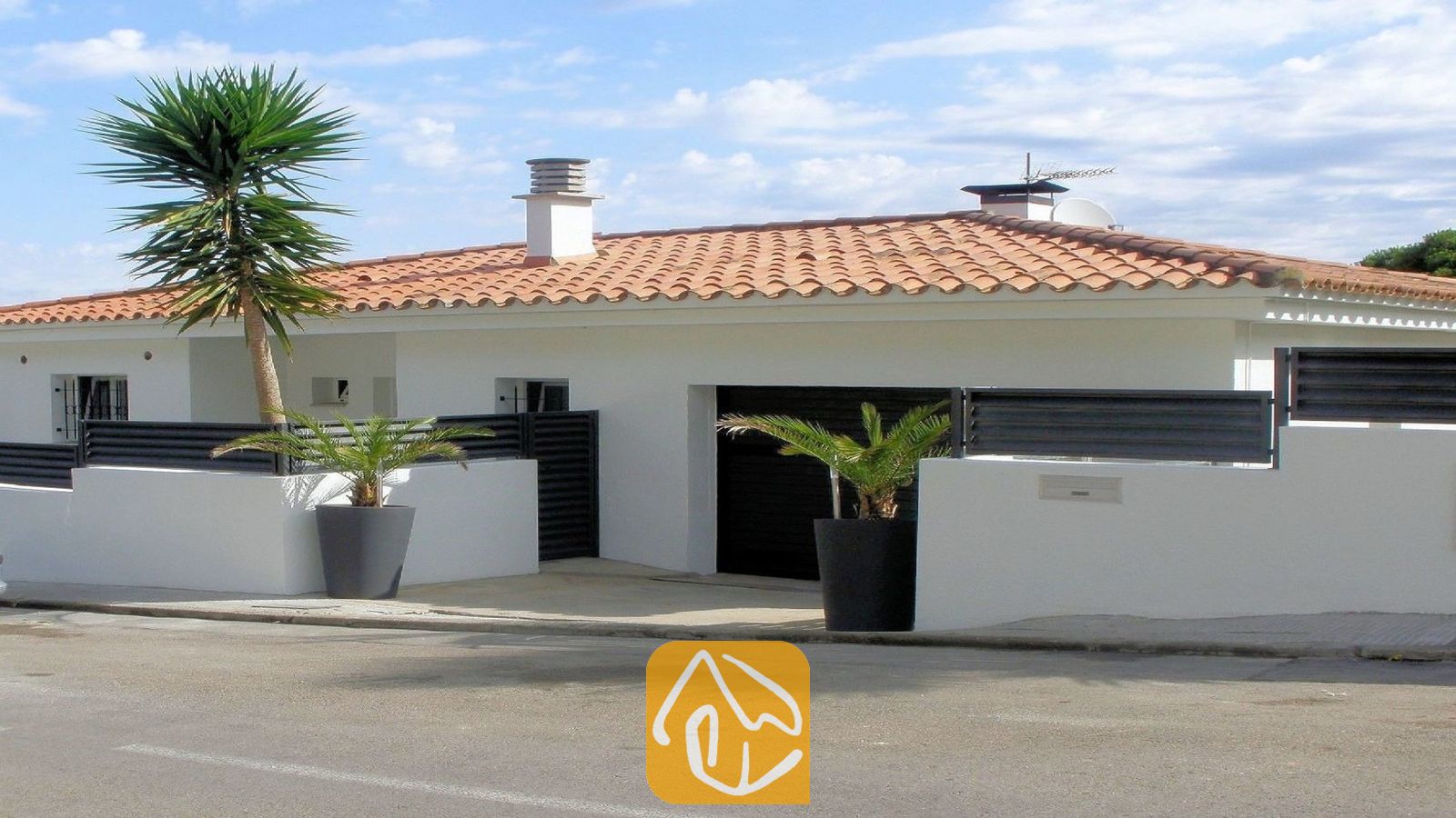 Vakantievilla's Costa Brava Spanje - Villa Fransisca - Buitenaanzicht van Villa Fransisca met witte muren, een oranje pannendak, een donkere garagedeur en potten met palmbomen.