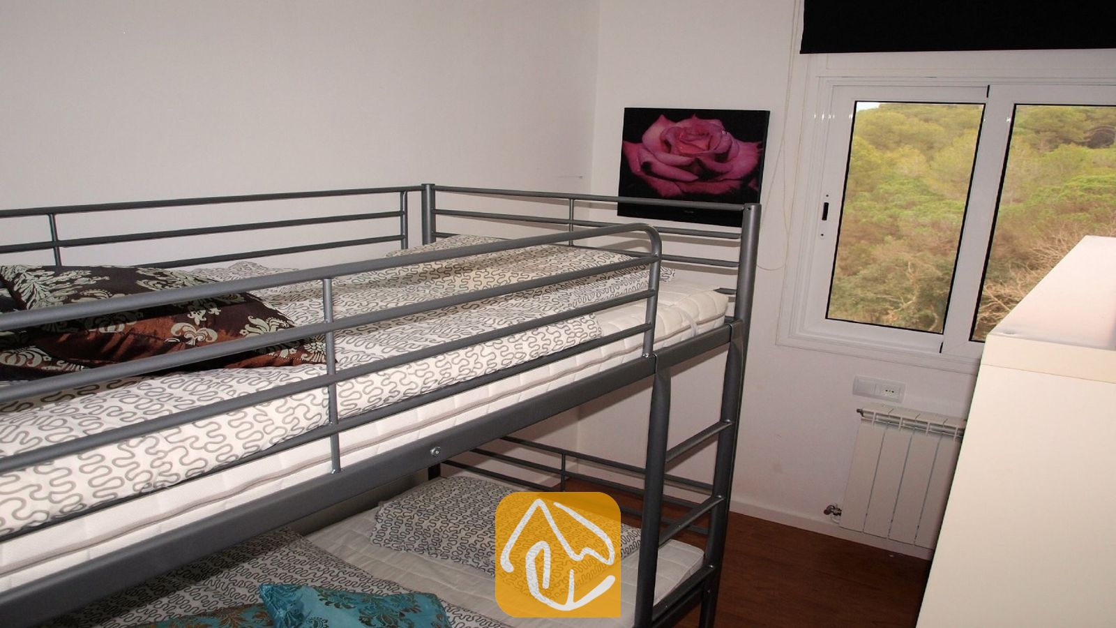 Vakantievilla's Costa Brava Spanje - Villa Fransisca - Slaapkamer met grijs metalen stapelbed, beddengoed met patroon, een roze wanddecoratie met een roos en een raam met uitzicht op de bomen.