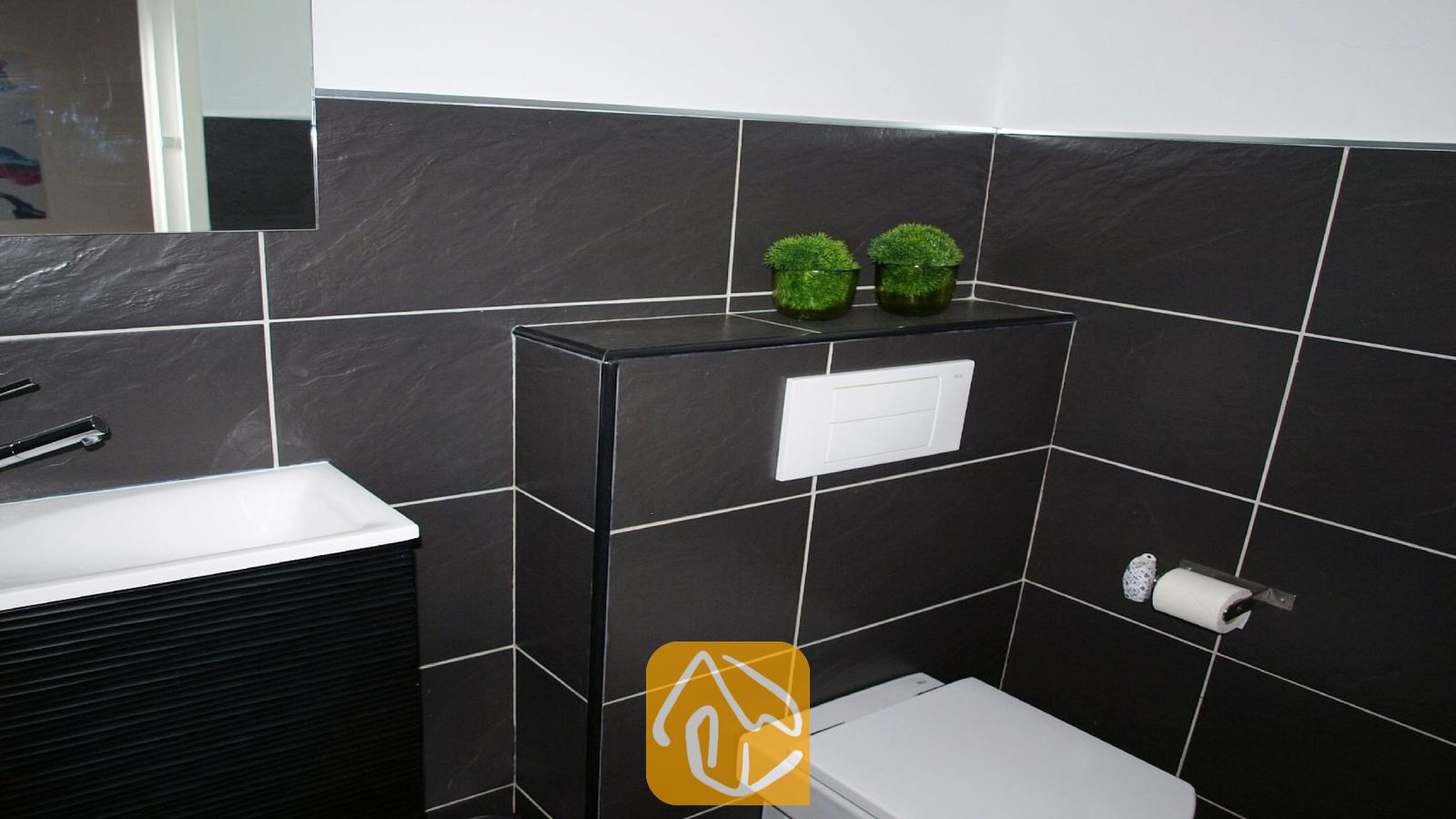 Vakantievilla's Costa Brava Spanje - Villa Fransisca - Moderne badkamer met donkergrijze vierkante wandtegels, een witte wastafel met een zwart badmeubel en een hangend toilet met daarboven twee kleine groene planten in pot.