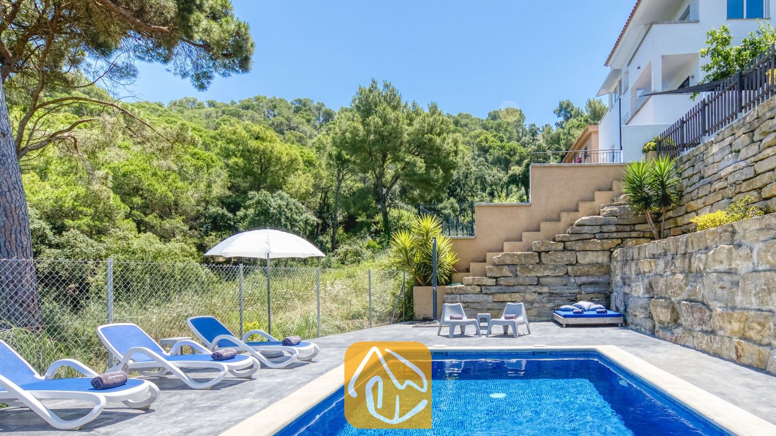 Vakantievilla's Costa Brava Spanje - Villa Fransisca - Privézwembad bij Villa Fransisca, met helderblauw water, witte ligbedden en een omringende stenen keermuur tegen de achtergrond van een heuvelachtig bos
