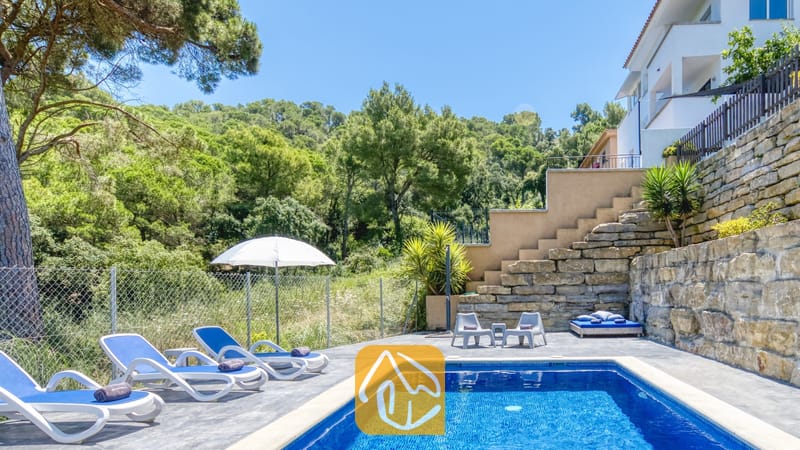 Vakantievilla's Costa Brava Spanje - Villa Fransisca - Privézwembad bij Villa Fransisca, met helderblauw water, witte ligbedden en een omringende stenen keermuur tegen de achtergrond van een heuvelachtig bos