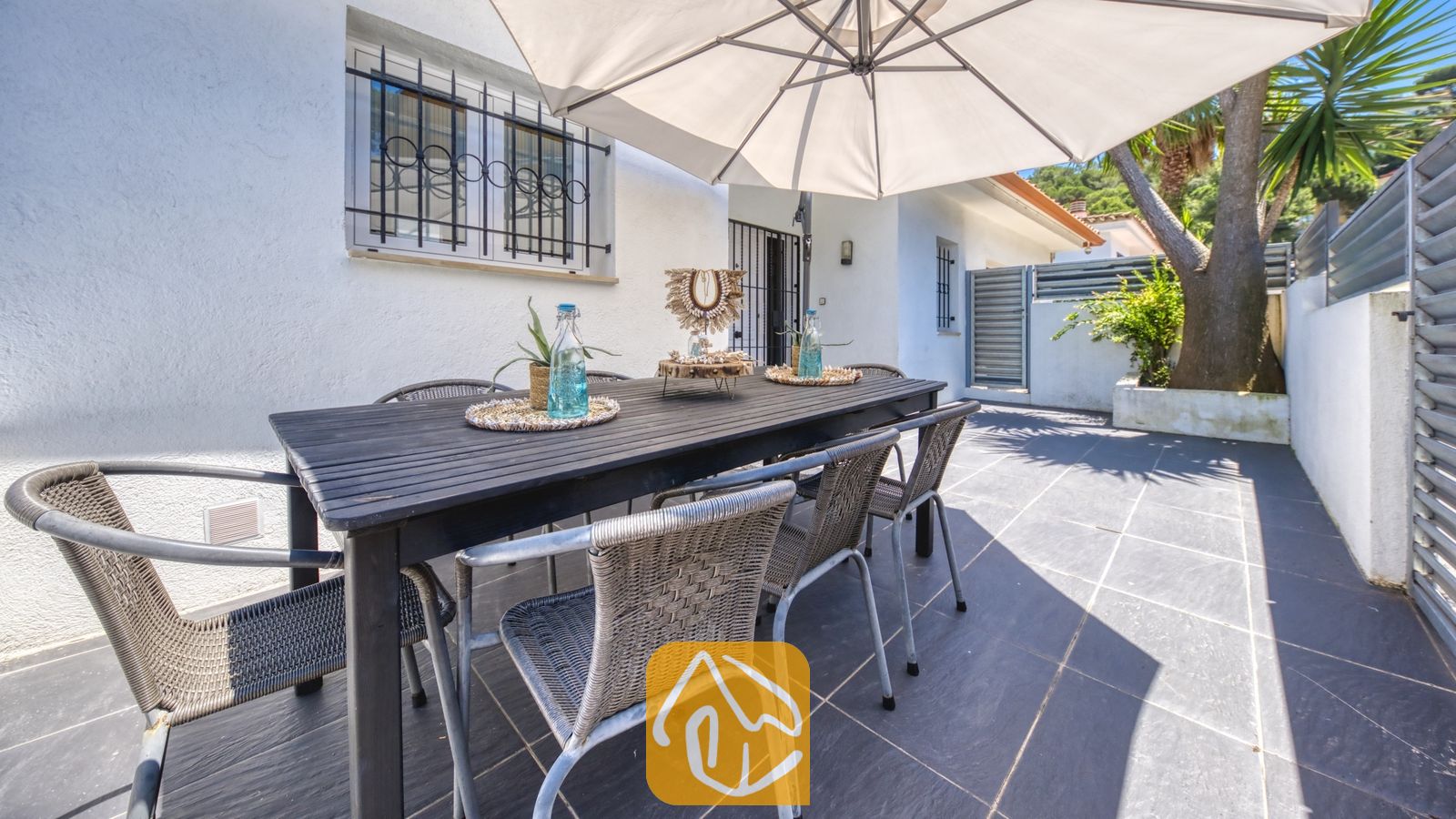 Vakantievilla's Costa Brava Spanje - Villa Fransisca - Buiten-eethoek bij Villa Fransisca met een donkerhouten tafel, omringd door geweven grijskleurige stoelen onder een grote witte parasol op een terras met tegels.