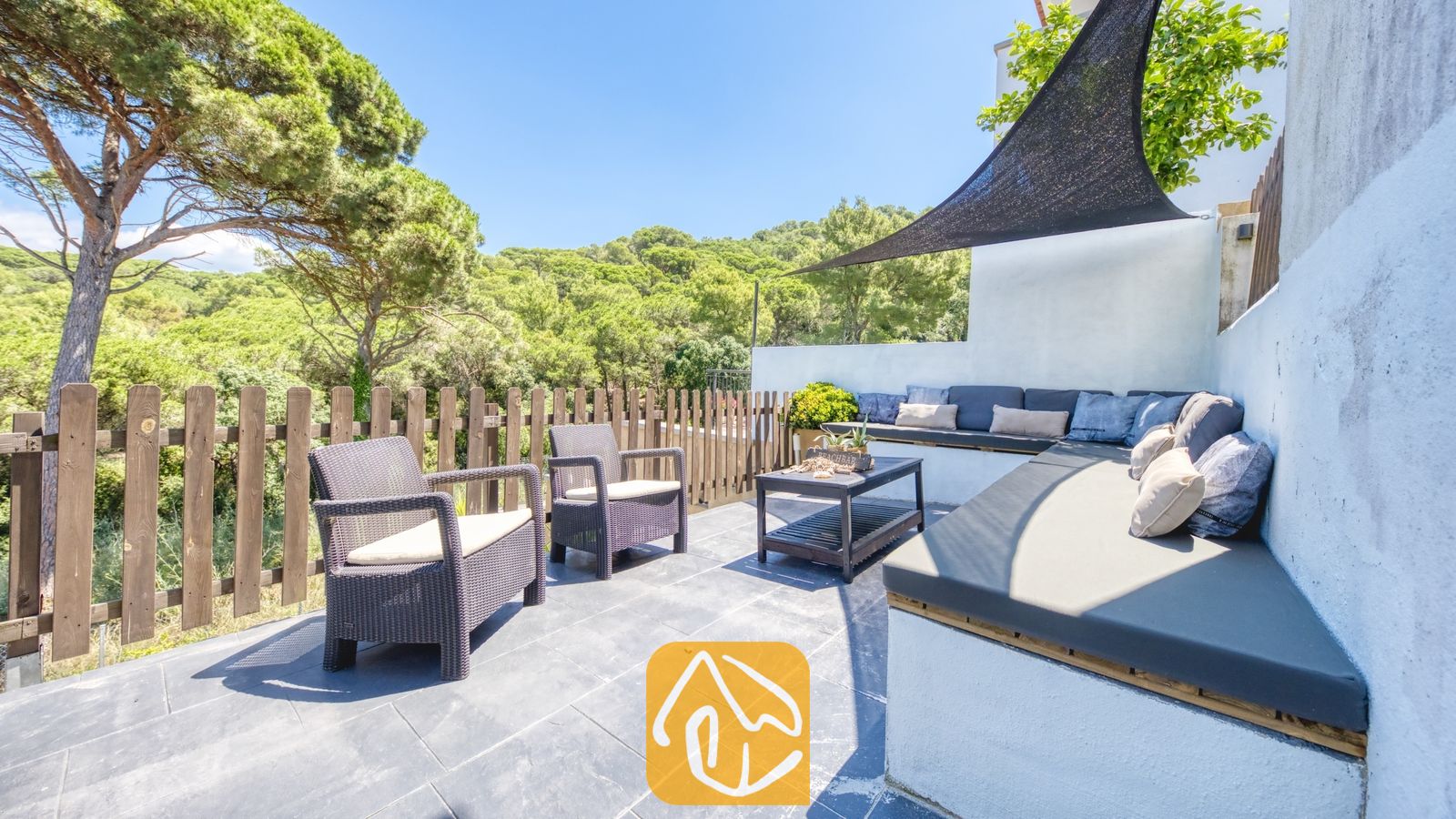 Vakantievilla's Costa Brava Spanje - Villa Fransisca - Terras bij Villa Fransisca met donkerwicker fauteuils, een ingebouwde witte zitbank met grijze kussens, een houten salontafel en een zwarte schaduwdoek met uitzicht op dennenbomen.