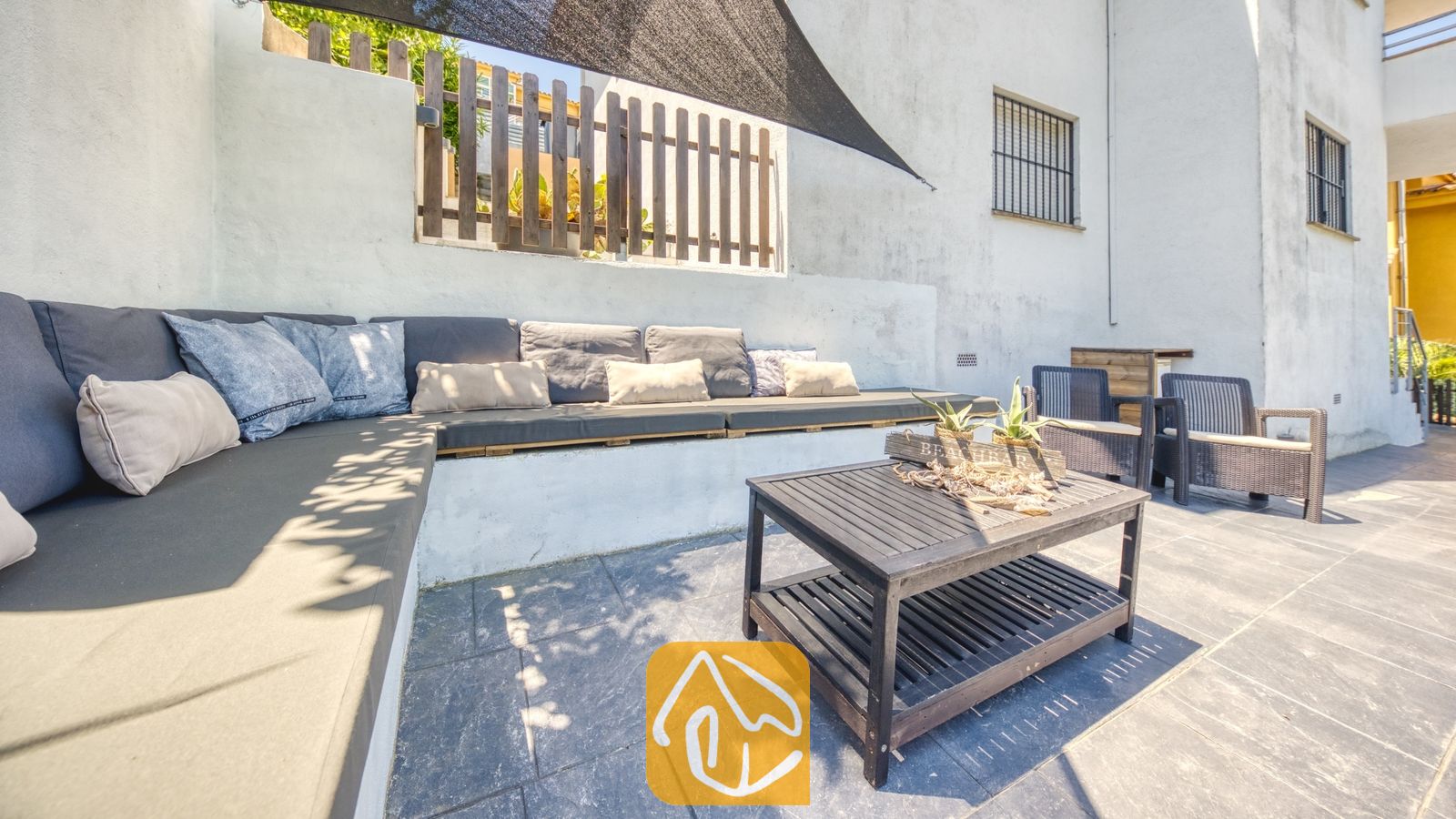 Vakantievilla's Costa Brava Spanje - Villa Fransisca - Buitenlounge op het terras van Villa Fransisca, voorzien van een ingebouwde hoekbank met donkergrijze kussens en een donkerhouten salontafel op een tegelvloer.