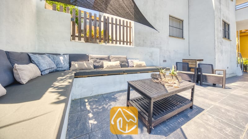 Vakantievilla's Costa Brava Spanje - Villa Fransisca - Buitenlounge op het terras van Villa Fransisca, voorzien van een ingebouwde hoekbank met donkergrijze kussens en een donkerhouten salontafel op een tegelvloer.