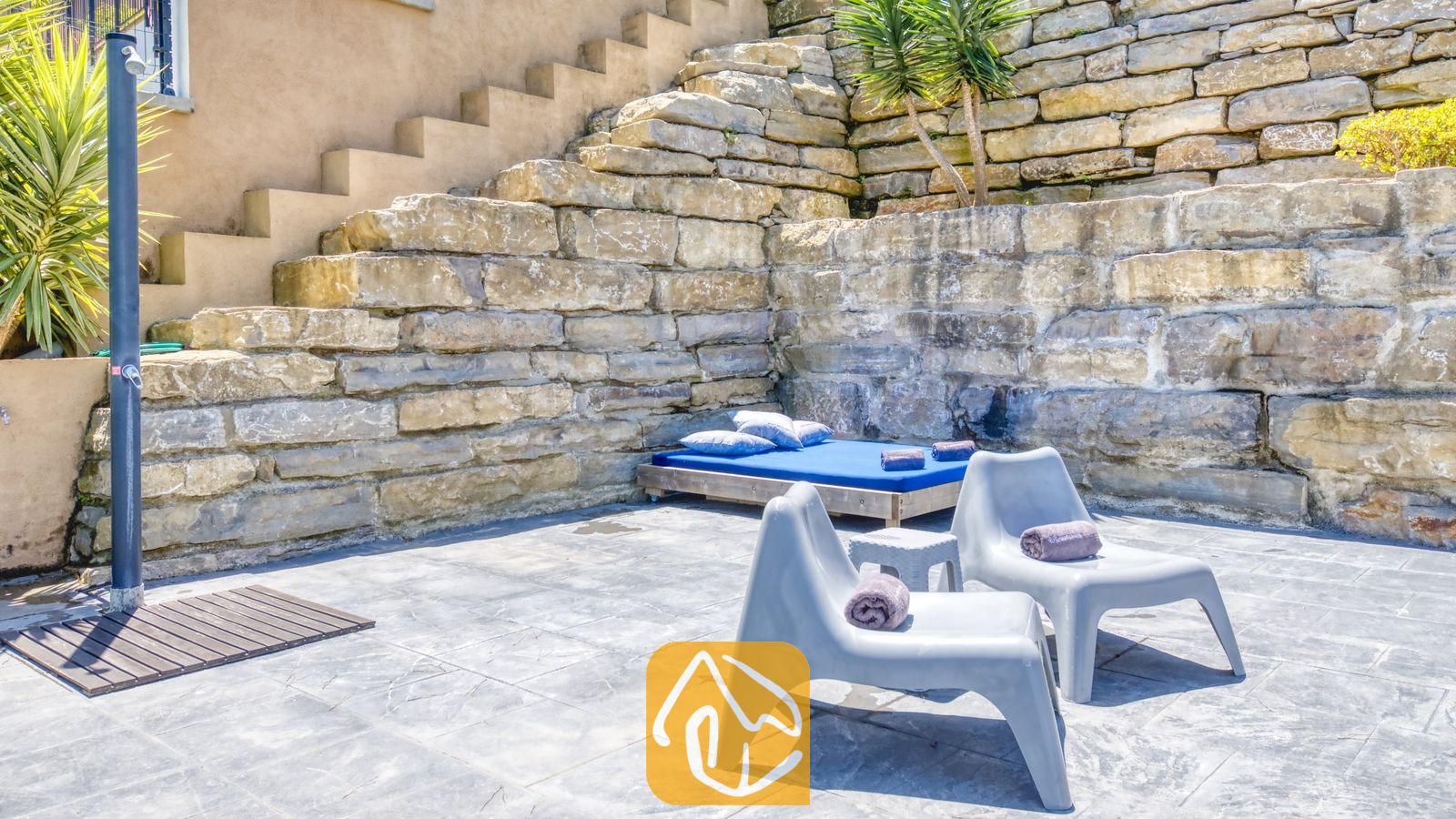Vakantievilla's Costa Brava Spanje - Villa Fransisca - Buitenpatio van Villa Fransisca met grijze kunststof ligstoelen, een blauw dagbed en een staande douche tegen een rustieke stenen keermuur.