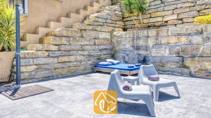 Vakantievilla's Costa Brava Spanje - Villa Fransisca - Buitenpatio van Villa Fransisca met grijze kunststof ligstoelen, een blauw dagbed en een staande douche tegen een rustieke stenen keermuur.