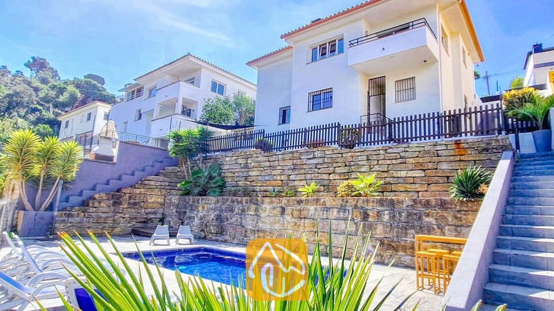 Vakantievilla's Costa Brava Spanje - Villa Fransisca - Buitenaanzicht van Villa Fransisca met een rechthoekig zwembad tegen getrapte stenen keermuren, onder een wit gebouw van meerdere verdiepingen en een blauwe hemel.