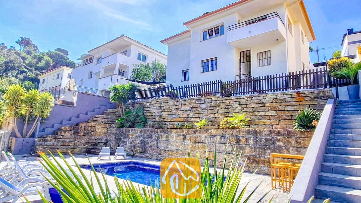 Vakantievilla's Costa Brava Spanje - Villa Fransisca - Buitenaanzicht van Villa Fransisca met een rechthoekig zwembad tegen getrapte stenen keermuren, onder een wit gebouw van meerdere verdiepingen en een blauwe hemel.