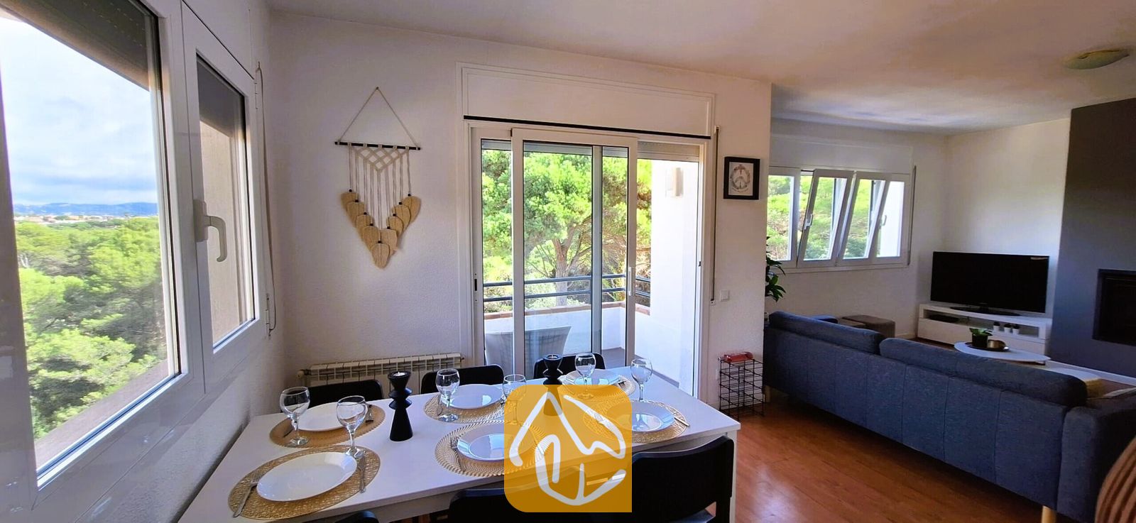 Vakantievilla's Costa Brava Spanje - Villa Fransisca - Eethoek binnen in Villa Fransisca, voorzien van een witte tafel gedekt voor zes personen, zwarte stoelen, geweven placemats en grote ramen met uitzicht op groene bomen.