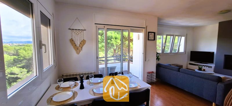 Villas de vacances Costa Brava Espagne - Villa Fransisca - Espace salle à manger à l'intérieur de la Villa Fransisca, doté d'une table blanche dressée pour six personnes, de chaises noires, de sets de table tissés et de grandes baies vitrées offrant une vue sur les arbres verdoyants.
