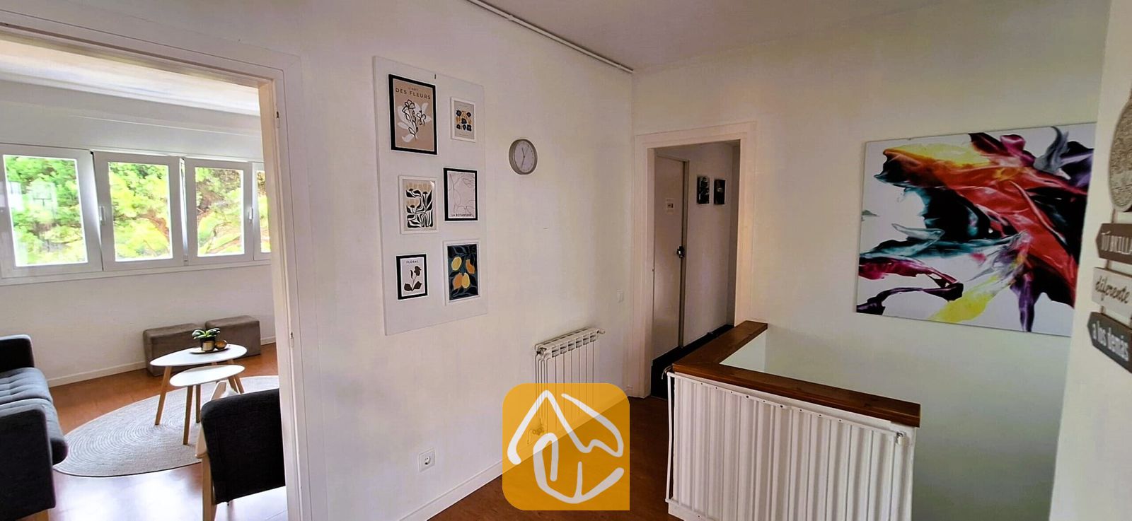 Vakantievilla's Costa Brava Spanje - Villa Fransisca - Gang in Villa Fransisca, voorzien van witte muren, houten vloeren, ingelijste botanische kunstwerken en een deuropening die leidt naar een verlichte kamer met een salontafel en een raam.
