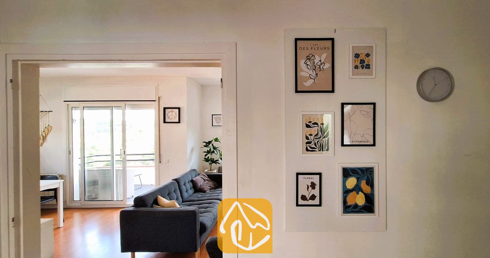 Vakantievilla's Costa Brava Spanje - Villa Fransisca - Uitzicht op de hal met een galerijwand van ingelijste botanische kunstprints en een ronde wandklok, die leidt naar een woonkamer met een donkere bank.