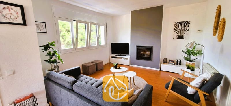 Villas de vacances Costa Brava Espagne - Villa Fransisca - Salon doté d'un canapé gris foncé, de parquet au sol, d'une cheminée encastrée, d'une télévision et de fenêtres blanches offrant une vue sur la verdure.