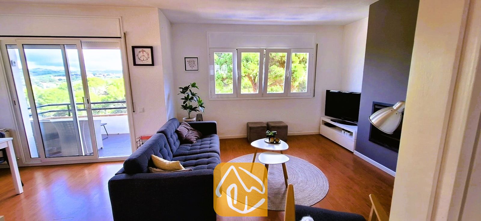 Vakantievilla's Costa Brava Spanje - Villa Fransisca - Woonkamer met een gecapitonneerde donkergrijze bank, in elkaar schuivende witte salontafels op een rond geweven vloerkleed, en een schuifpui die toegang biedt tot een balkon.