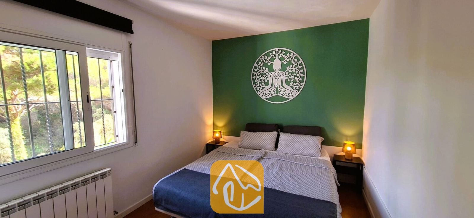 Vakantievilla's Costa Brava Spanje - Villa Fransisca - Slaapkamer met een tweepersoonsbed, gedessineerd grijs beddengoed en twee kleine nachtlampjes tegen een groene accentmuur met een witte, ronde boomsculptuur.