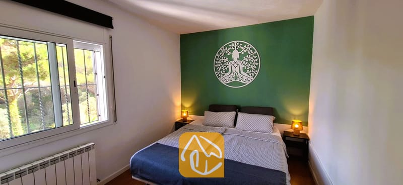 Villas de vacances Costa Brava Espagne - Villa Fransisca - Chambre avec un lit double, du linge de lit gris à motifs et deux petites lampes de chevet, disposés contre un mur d'accent vert agrémenté d'une sculpture d'arbre circulaire blanche.
