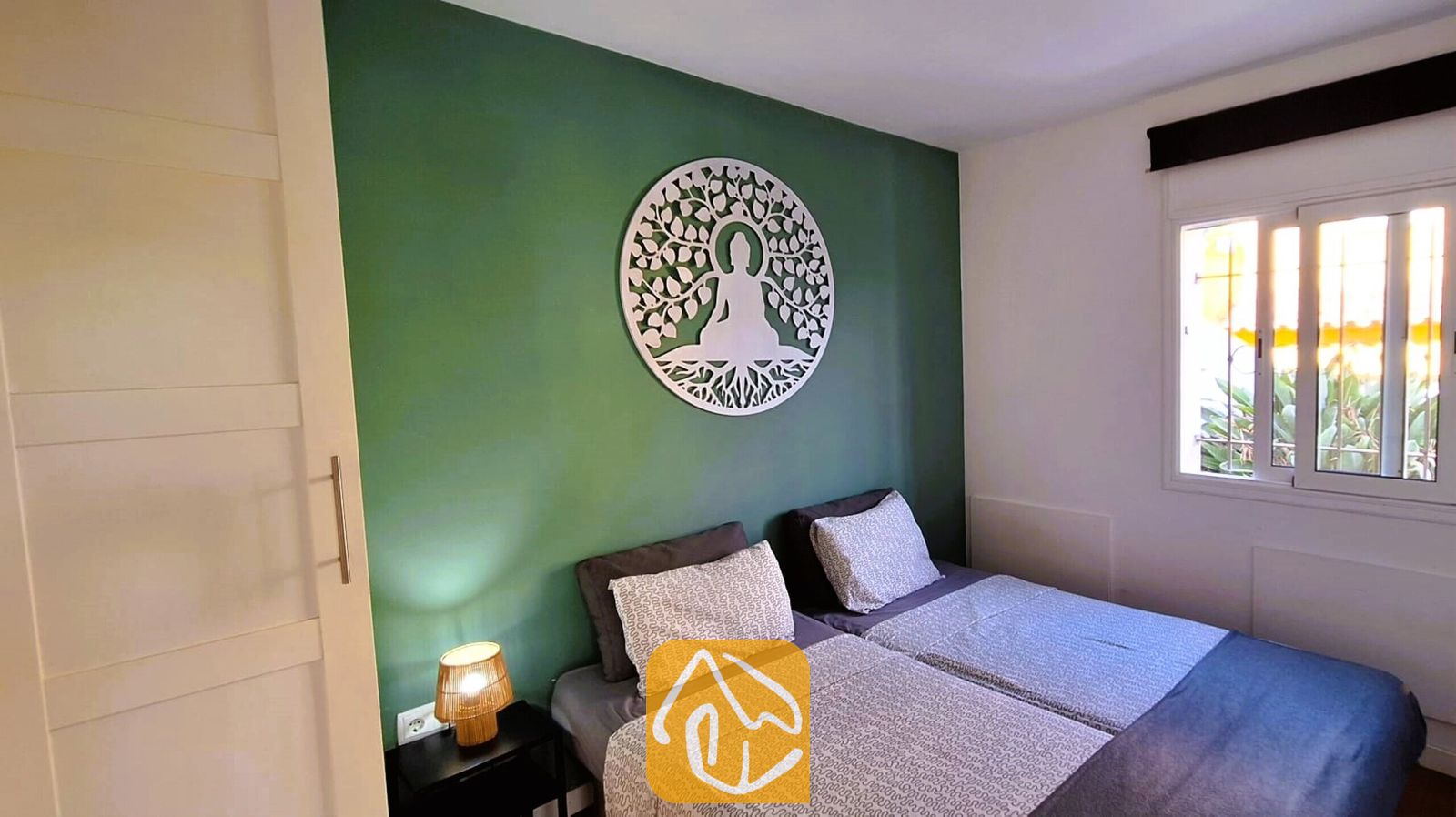 Vakantievilla's Costa Brava Spanje - Villa Fransisca - Slaapkamer met twee eenpersoonsbedden tegen een groene wand, voorzien van een groot rond, wit wandobject en een nachtkastlampje.