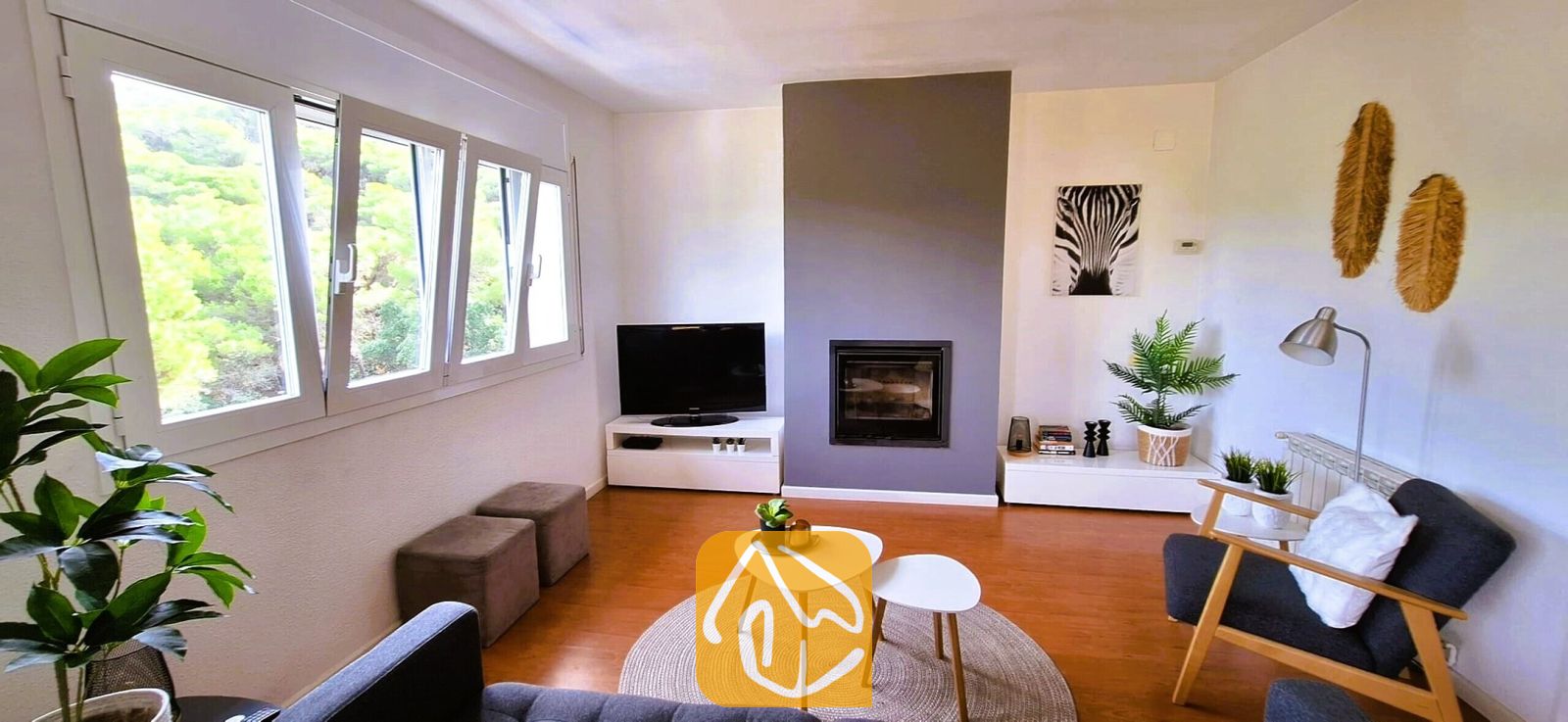 Vakantievilla's Costa Brava Spanje - Villa Fransisca - Woonkamer met een ingebouwde open haard tegen een grijze accentmuur, twee witte in elkaar schuivende salontafels op een rond geweven vloerkleed en een televisie op een wit televisiemeubel.