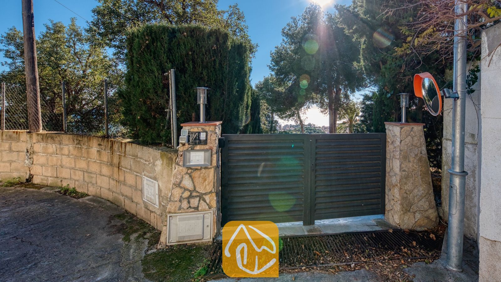 Vakantievilla's Costa Brava Spanje - Villa Gabriella - Toegangspoort van Villa Gabriella met donkergrijze horizontale lamellen, stenen zuilen met verlichting en een beveiligingspaneel, omlijst door een achtergrond van hoge bomen.
