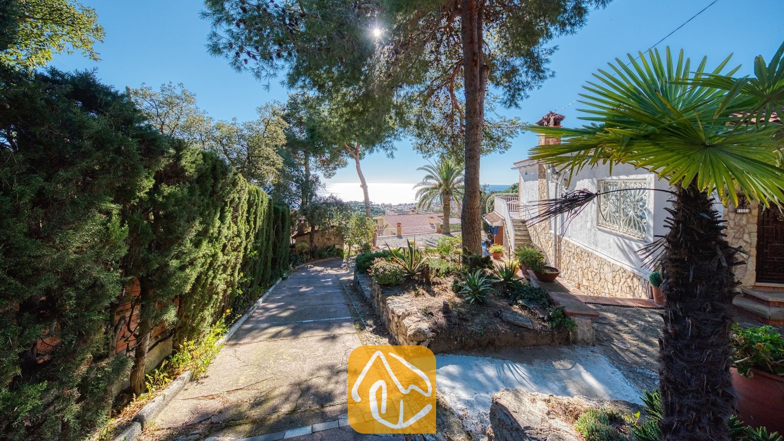 Vakantievilla's Costa Brava Spanje - Villa Gabriella - De verharde oprijlaan van Villa Gabriella, geflankeerd door hoge groene heggen en stenen plantenbakken, leidt naar een prachtig uitzicht op de zee onder een stralend blauwe lucht.