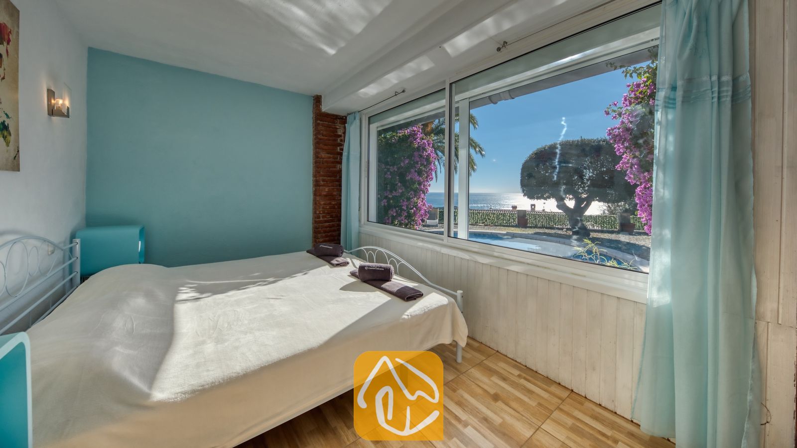 Vakantievilla's Costa Brava Spanje - Villa Gabriella - Slaapkamer met een wit metalen bed en een groot raam met uitzicht op de zee en het zwembad. De kamer is voorzien van een lichtblauwe accentmuur, een vloer met houtlook en paarse accenten.