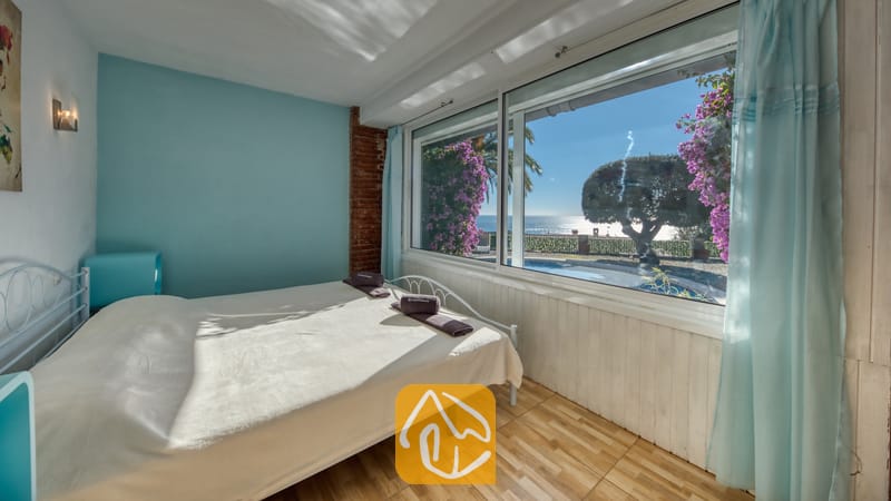 Vakantievilla's Costa Brava Spanje - Villa Gabriella - Slaapkamer met een wit metalen bed en een groot raam met uitzicht op de zee en het zwembad. De kamer is voorzien van een lichtblauwe accentmuur, een vloer met houtlook en paarse accenten.